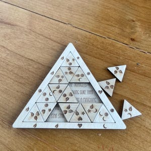 May include: A wooden puzzle in the shape of a large triangle, with smaller triangular pieces. The puzzle has engraved floral and celestial designs. Text on the puzzle reads "THE TRIANGLES MAKING SURE THAT A MATCHING STAR STAR".