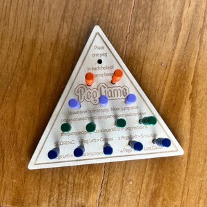 May include: A wooden triangular game board with the words "Peg Game" printed on it. The board has 15 holes, each with a peg. The pegs are orange, green, and blue. The board also has instructions for playing the game, including scoring.