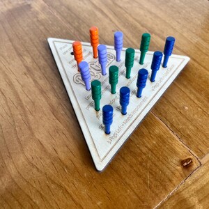May include: A wooden triangular game board with the word "Peg-a-Way" printed on it. The board has 15 holes with pegs in them. The pegs are in different colors: orange, blue, and green. The board has instructions printed on it, including "3 Pegs Left = Needs Improvement" and "4 Pegs Left = Lost Cause".