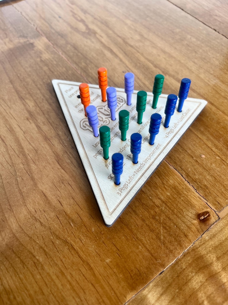 May include: A wooden triangular game board with the word "Peg-a-Way" printed on it. The board has 15 holes with pegs in them. The pegs are in different colors: orange, blue, and green. The board has instructions printed on it, including "3 Pegs Left = Needs Improvement" and "4 Pegs Left = Lost Cause".