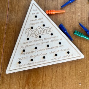May include: Wooden triangular game board with the text "Peg Game" and instructions for playing. The board has 15 holes and is designed for a peg game.