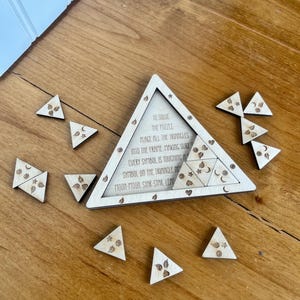 May include: Wooden triangle puzzle with a matching frame. The frame has text that reads "To solve the puzzle, place all the triangles into the frame..." The puzzle pieces are small triangles with etched designs of moons, stars, and leaves.