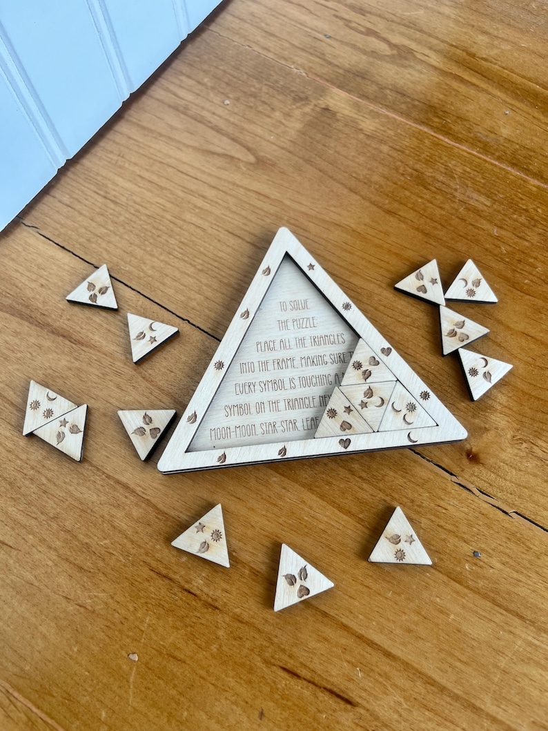May include: Wooden triangle puzzle with a matching frame. The frame has text that reads "To solve the puzzle, place all the triangles into the frame..." The puzzle pieces are small triangles with etched designs of moons, stars, and leaves.