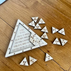 May include: Wooden triangle puzzle with engraved symbols and text. The puzzle includes a triangular frame and several smaller triangle pieces with leaf, moon, and star designs. The text reads "To solve the puzzle..." and provides instructions.