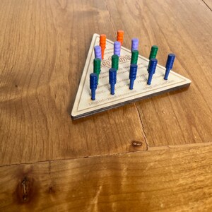 May include: A wooden triangle game board with pegs in various colors, including orange, purple, green, and blue. The board has text that reads "Pegs Left - Needs Improvement" and "Pegs Left - Lost Game".