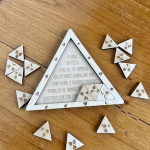 May include: A wooden puzzle featuring a triangular frame and numerous smaller triangular pieces. The frame is inscribed with text, including "TO SOLVE THE PUZZLE". The pieces are decorated with engraved symbols such as stars, moons, and leaves.