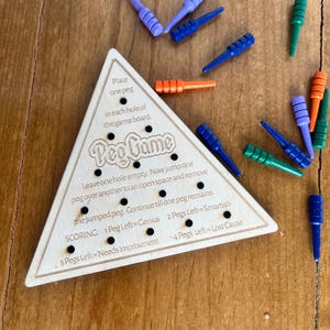 May include: A wooden triangular game board with the words "Peg Game" printed on it. The board has holes for pegs and instructions for playing the game. The game includes colorful pegs in blue, green, orange, and purple.