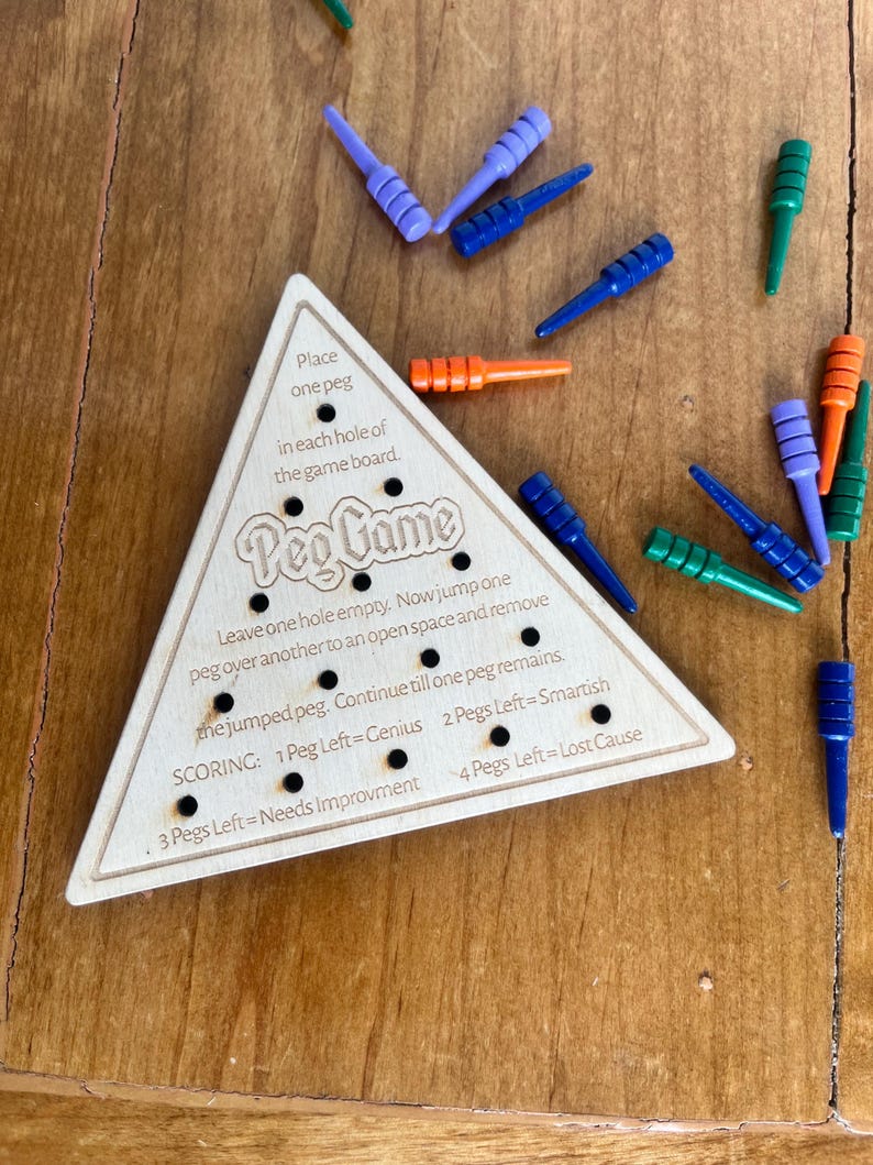 May include: A wooden triangular game board with the words "Peg Game" printed on it. The board has holes for pegs and instructions for playing the game. The game includes colorful pegs in blue, green, orange, and purple.