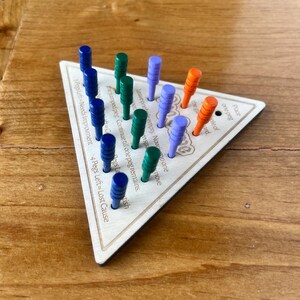 May include: A wooden triangular game board with pegs in various colors, including blue, green, purple, and orange. The board has text that reads "Leave one peg empty. Now move the jumped peg. Continue till one peg remains." and "Pegs Left=Needs Improvement 4 Pegs Left=Lost Cause".