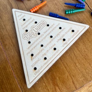 May include: A wooden triangular game board with holes and pegs. The board has the text "Peg Game" and instructions for playing the game. The text on the board reads: "Place one peg in each hole of the game board. Leave one hole empty. Now jump one peg over another to an open space and remove the jumped peg. Continue till one peg remains. SCORING: 1 Peg Left=Genius 2 Pegs Left=Smartish 3 Pegs Left = Needs Improvement 4 Pegs Left=Lost Cause"