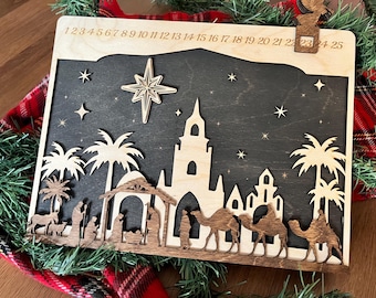May include: Wooden Christmas advent calendar with a laser-cut nativity scene. The design features a star, palm trees, and a church. Numbers 1-25 are etched across the top. The calendar is made of light and dark wood.