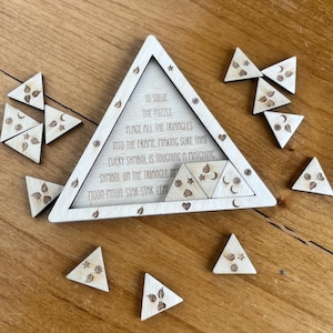 May include: Wooden triangle puzzle with engraved symbols and text. The puzzle includes a triangular frame and several smaller triangular pieces. The text reads, "To solve the puzzle, place all the triangles into the frame..." The symbols include moons, stars, and leaves.