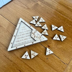 May include: A wooden triangle puzzle with a matching frame and several loose triangular pieces. The puzzle pieces and frame are engraved with celestial and botanical symbols. Text on the frame reads, "TO SOLVE THE PUZZLE..."