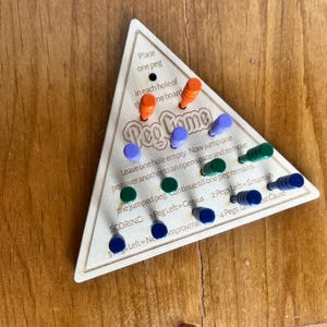 May include: Wooden triangular game board with the words "Peg Game" printed on it. The board has holes for pegs and instructions for playing the game. The game includes colorful pegs in blue, green, orange, and red.