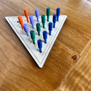 May include: A wooden triangular game board with pegs in various colors, including blue, green, orange, and purple. The board has text that reads "Place one peg near the... Leave peg... more to the... Score 3 Pegs Left=Needs Improvement 4 Pegs Left=Must Continue".