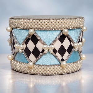 May include: A cylindrical decorative box featuring a light blue velvet finish and a black and white diamond pattern. The exterior is adorned with silver braided trim, rows of small white pearls, and larger pearl accents shaped like bows.