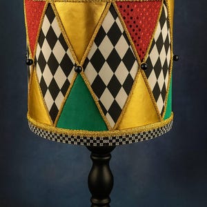 May include: A table lamp featuring a cylindrical shade with alternating panels of black and white diamonds, solid gold, solid green, and red glitter fabric. The shade is trimmed with gold cord, black beads, and a black and white checkered border.