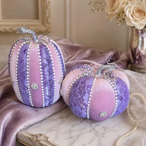 May include: Two decorative pumpkins with a velvet and lace design. The pumpkins are in shades of purple and pink, adorned with pearl accents and sparkling embellishments. The stems are silver. These pumpkins are perfect for fall decor.