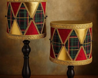 May include: Two decorative drum-shaped lamps with black bases. The lampshades feature a red fabric with gold accents and a green and black plaid pattern. The lamps are on a wooden surface.
