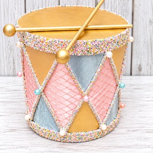 May include: A decorative drum featuring a gold top surface with two gold drumsticks resting on it. The sides are adorned with a diamond pattern in pink, blue, and gold, accented by colorful sprinkles along the edges and small pearl-like beads.