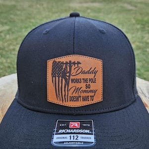 Può includere: Cappellino da camionista nero con retro in rete e toppa in similpelle marrone. La toppa presenta il testo "Daddy Works The Pole So Mommy Doesn't Have To" e un'illustrazione di un elettricista e della bandiera americana.