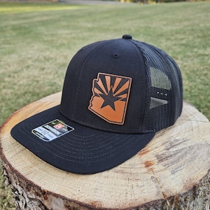 May include: A black and brown trucker hat with a leather patch featuring the outline of Arizona with a star and sun rays. The hat has a black mesh back.
