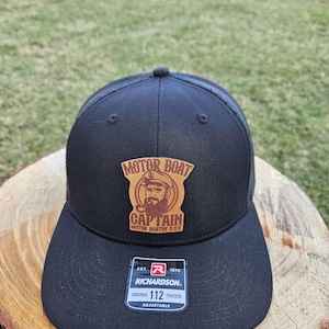 May include: Black trucker hat with a curved peak and mesh back. The hat features a leather patch with the words "MOTOR BOAT CAPTAIN" and a graphic of a bearded man. The hat also has a "RICHARDSON" tag.