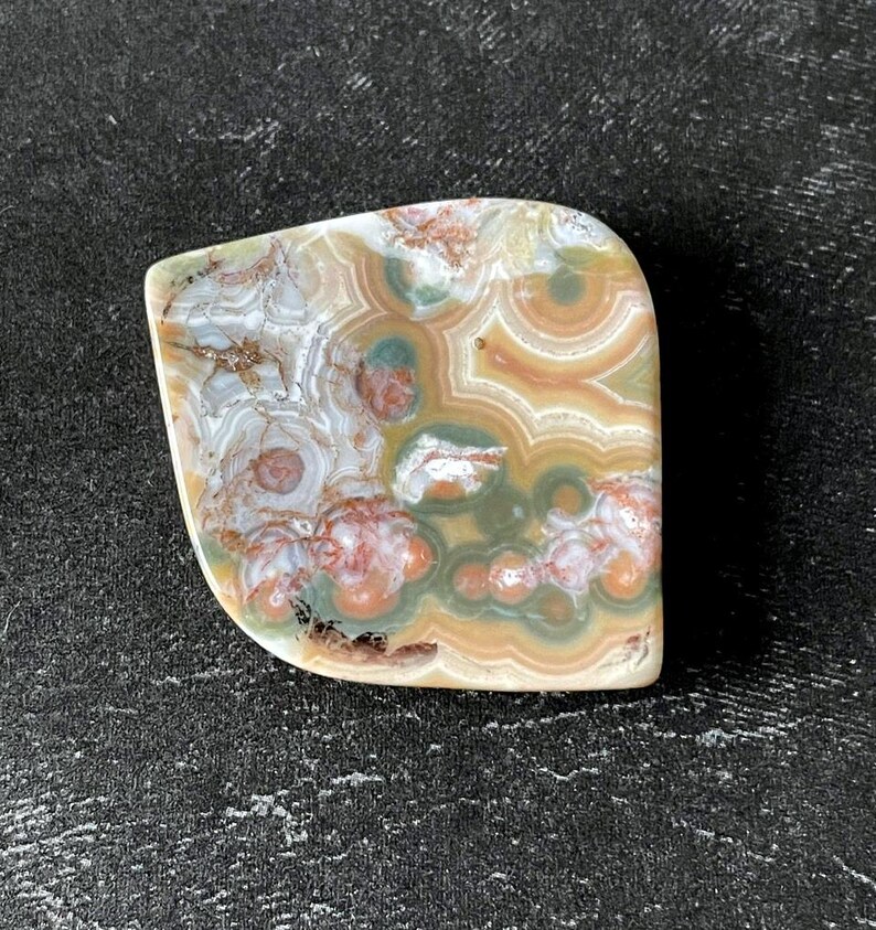 May include: A polished, irregularly shaped stone with a unique pattern. The stone features swirls of white, brown, green, and reddish-orange hues. The surface has a smooth, glossy finish, and the stone is set against a dark background.
