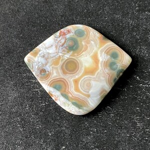 May include: A polished, rhomboid-shaped gemstone with a unique pattern. The stone features concentric circles in shades of tan, green, and white, with hints of red. The overall design resembles a natural, abstract landscape.