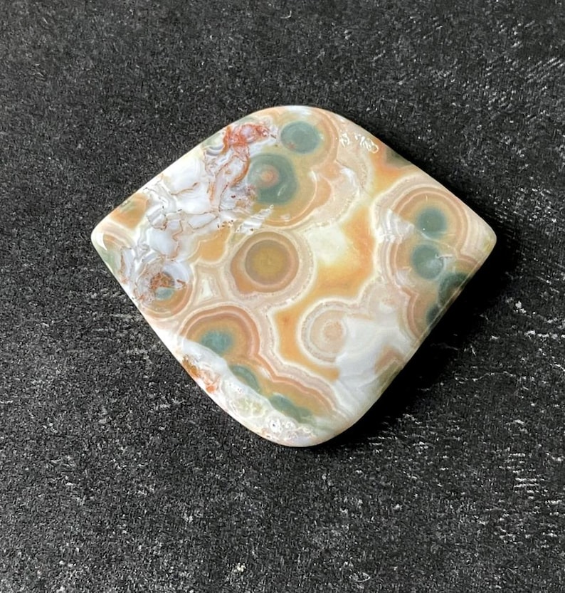 May include: A polished, rhomboid-shaped gemstone with a unique pattern. The stone features concentric circles in shades of tan, green, and white, with hints of red. The overall design resembles a natural, abstract landscape.
