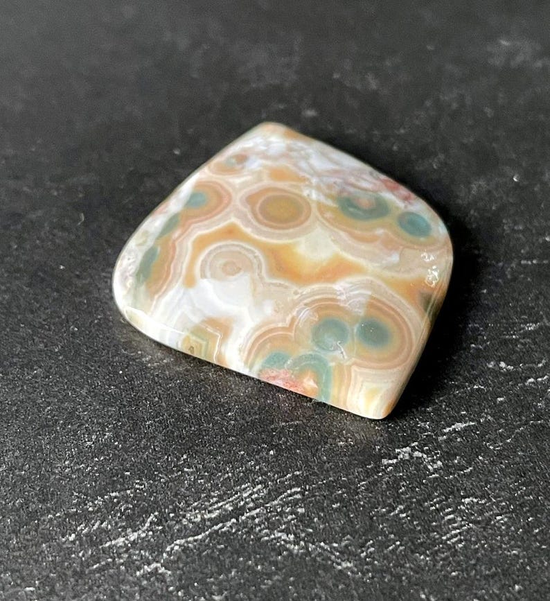May include: A polished gemstone with a unique pattern of concentric circles in shades of beige, brown, green, and white. The stone has a smooth, rounded shape and is set against a dark background.
