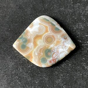 May include: A polished gemstone with a unique pattern of concentric circles in shades of beige, brown, green, and white. The stone is diamond-shaped with rounded corners, showcasing natural variations in color and texture.
