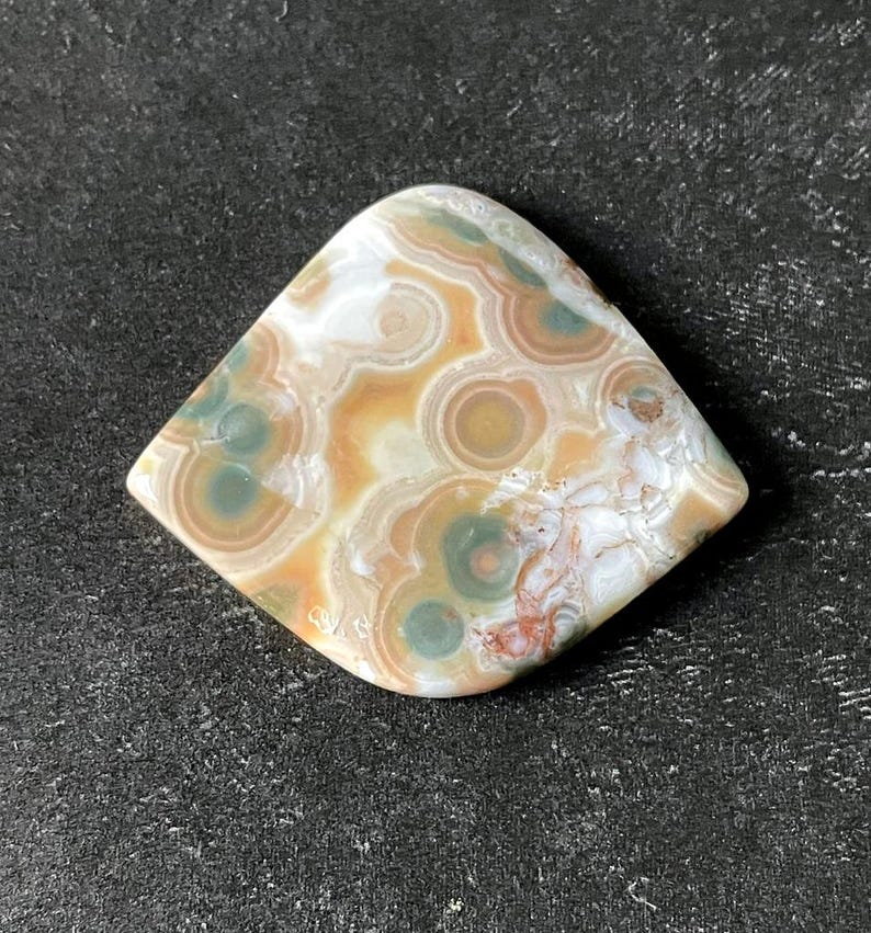 May include: A polished gemstone with a unique pattern of concentric circles in shades of beige, brown, green, and white. The stone is diamond-shaped with rounded corners, showcasing natural variations in color and texture.