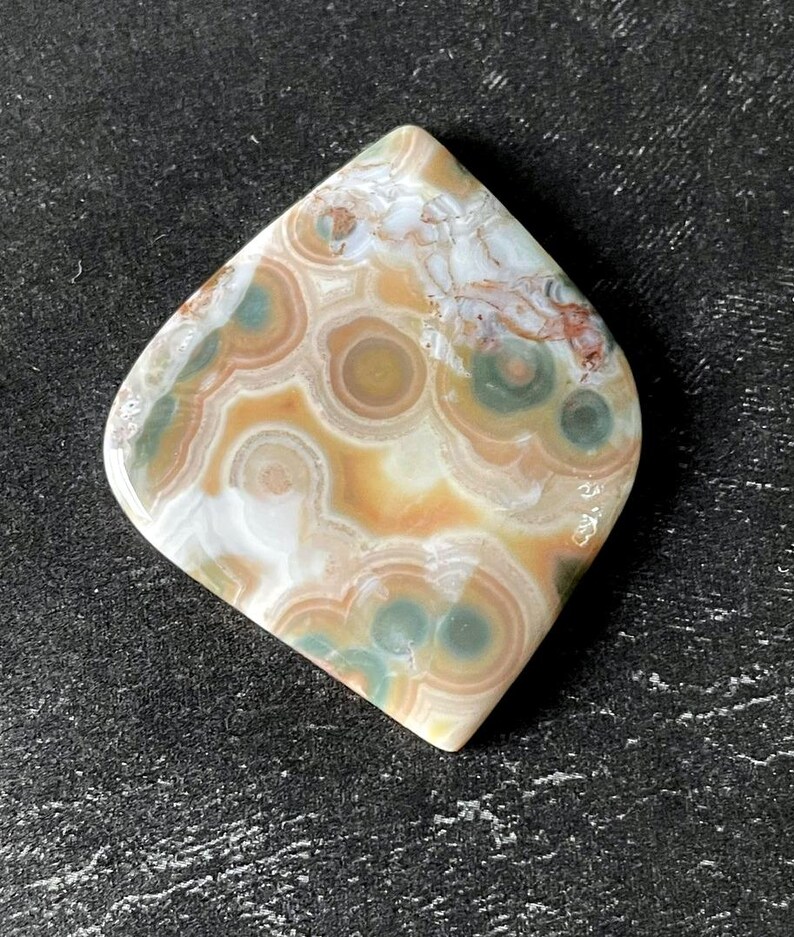 May include: A polished gemstone with a unique pattern of concentric circles in shades of brown, green, and white. The stone has a smooth, rounded shape and is set against a dark background.