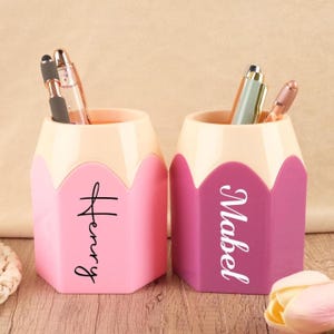 May include: Two pencil-shaped pen holders, one pink and one purple, each with a name in script. The pink holder says "Henry" and the purple holder says "Mabel". Each holder contains pens and pencils. The top of each holder is a light beige color.