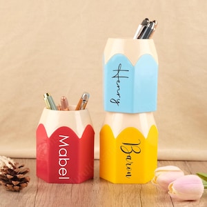 Könnte beinhalten: Drei stiftförmige Schreibtisch-Organizer in Rot, Blau und Gelb, jeweils mit weißem Oberteil und den Namen "Mabel", "Henry" und "Baren" bedruckt. Die Organizer halten Stifte und Bleistifte.
