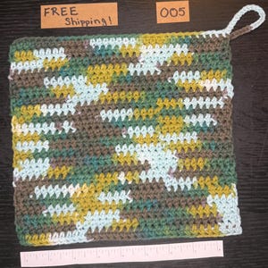 May include: A square, handmade crochet potholder in a camouflage pattern. The potholder has a loop for hanging and is made with green, brown, yellow, and light blue yarn. The potholder is approximately 20 cm square.