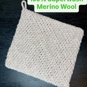 Puede incluir: Un artículo cuadrado de ganchillo beige claro con un lazo en una esquina. El artículo está hecho de un hilo texturizado y presenta el texto "100% Superwash Merino Wool" en un cuadro verde.