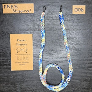 May include: A multi-coloured glasses chain with blue, yellow, and white woven threads. The chain has black rubber loops on each end. A small brown tag reads "Peeper Keepers" with the brand name "kuke" and social media handles. Another tag says "006".