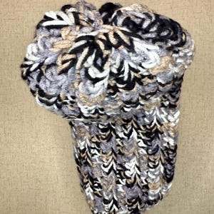 May include: A hand-knitted hat in a mix of black, white, grey, and tan yarns. The hat has a textured, patterned design. It is displayed on a black stand against a neutral background. The hat's design is intricate and visually appealing.