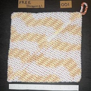 Puede incluir: Paño de cocina de ganchillo hecho a mano con un patrón de rayas diagonales en blanco, amarillo y naranja. El paño de cocina tiene un lazo para colgar y mide aproximadamente 20 cm cuadrados. El artículo se muestra sobre una superficie oscura.