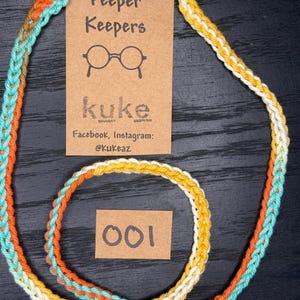 May include: A colorful eyeglass holder with a braided design in shades of orange, teal, and yellow. The holder features clear loops for attaching to glasses and a tag that reads "Peeper Keepers" with the brand name "Kuke" and social media handles. The number "001" is on a small tag.