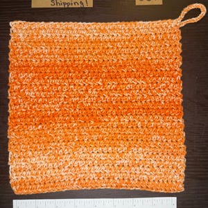 May include: Handmade orange and white crochet potholder with a loop for hanging. The square-shaped potholder features a textured stitch pattern. The colour gradient transitions from white to orange. A ruler is placed below the potholder.