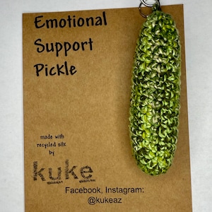 May include: A green and yellow crocheted pickle keyring with a silver clasp. The pickle is attached to a brown card with the text "Emotional Support Pickle" and "made with recycled silk by kuke".