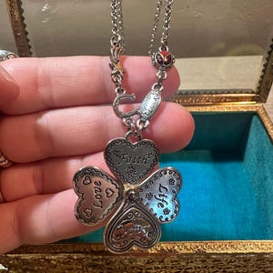 Pode incluir: Colar prateado com um pendente de trevo de quatro folhas. Cada folha em forma de coração é gravada com palavras como "Amor", "Fé" e "Vida". O colar inclui berloques com uma ferradura e uma estrela.