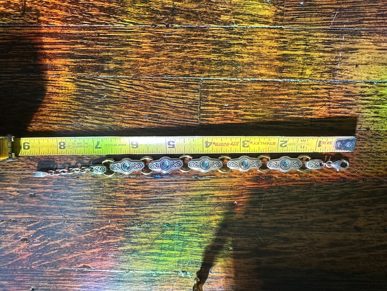 May include: Silver-toned bracelet with oval-shaped links, each featuring a central blue stone. The bracelet is approximately 6 inches long, as measured by a yellow tape measure. The bracelet has a decorative design.