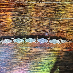 May include: Silver-toned bracelet with an ornate design. The bracelet features a series of oval-shaped links with circular and scroll details, connected by smaller links. The clasp is a lobster claw style.