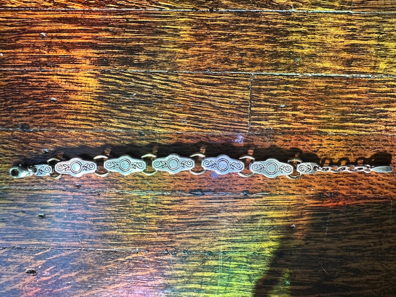 May include: Silver-toned bracelet with an ornate design. The bracelet features a series of oval-shaped links with circular and scroll details, connected by smaller links. The clasp is a lobster claw style.