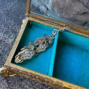 May include: A gold-toned, ornate jewelry box with a glass top, lined with teal velvet. Inside, a silver and gold-toned chain-linked bracelet with decorative circular and swirl designs rests. The box is open, revealing its contents.