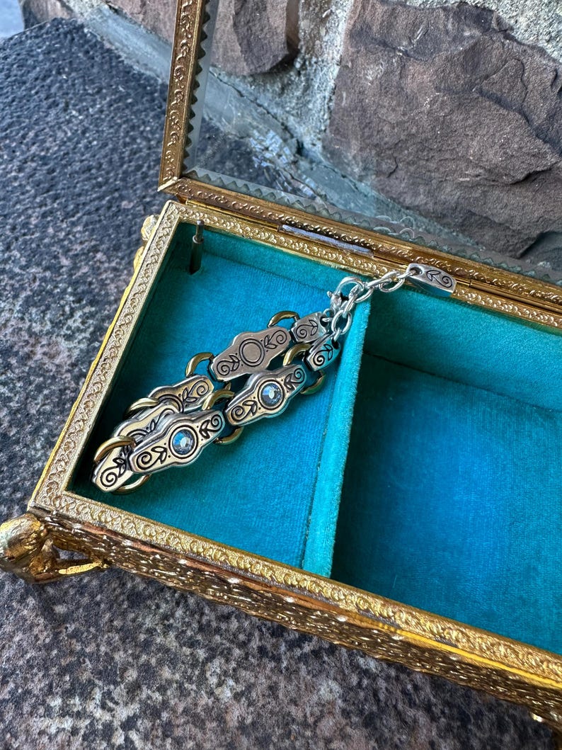 May include: A gold-toned, ornate jewelry box with a glass top, lined with teal velvet. Inside, a silver and gold-toned chain-linked bracelet with decorative circular and swirl designs rests. The box is open, revealing its contents.
